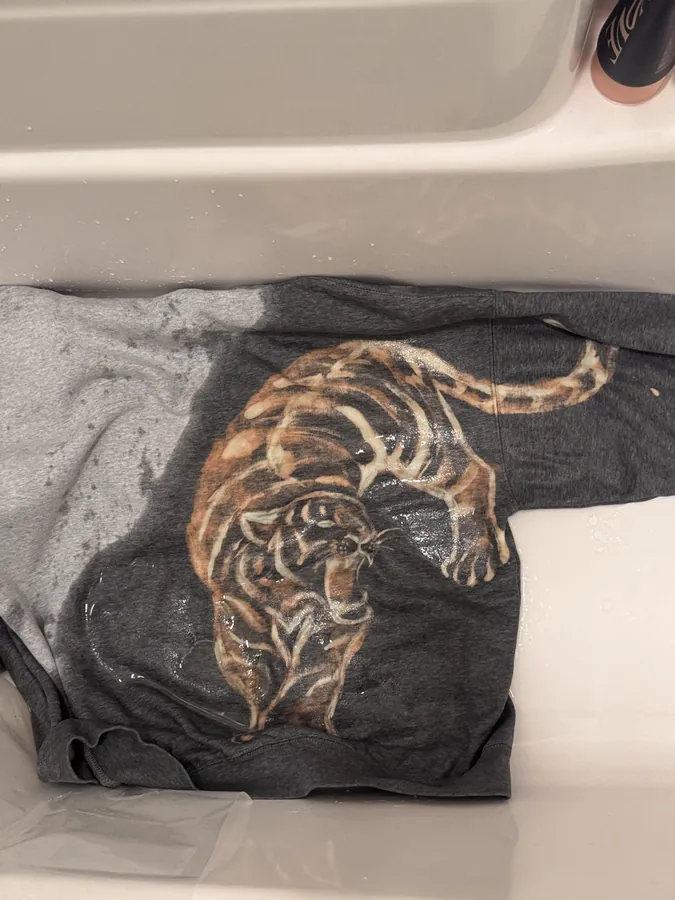 A dark grey hoodie with a crouching tiger painted onto it in bleach, laid wet in a basin.