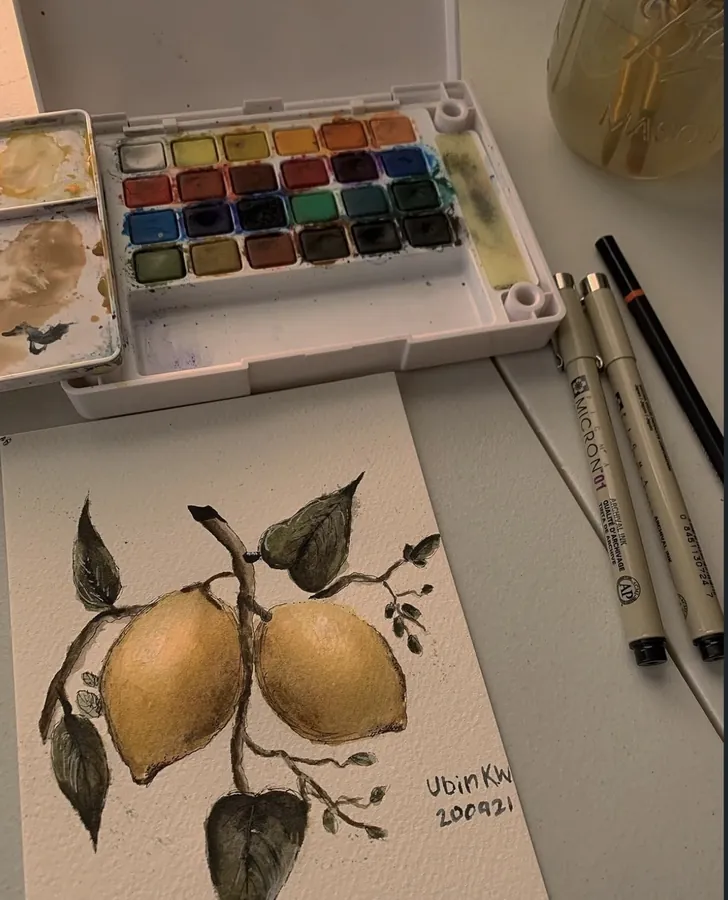 A watercolour painting of two lemons on a branch, beside a paint palette and pens.