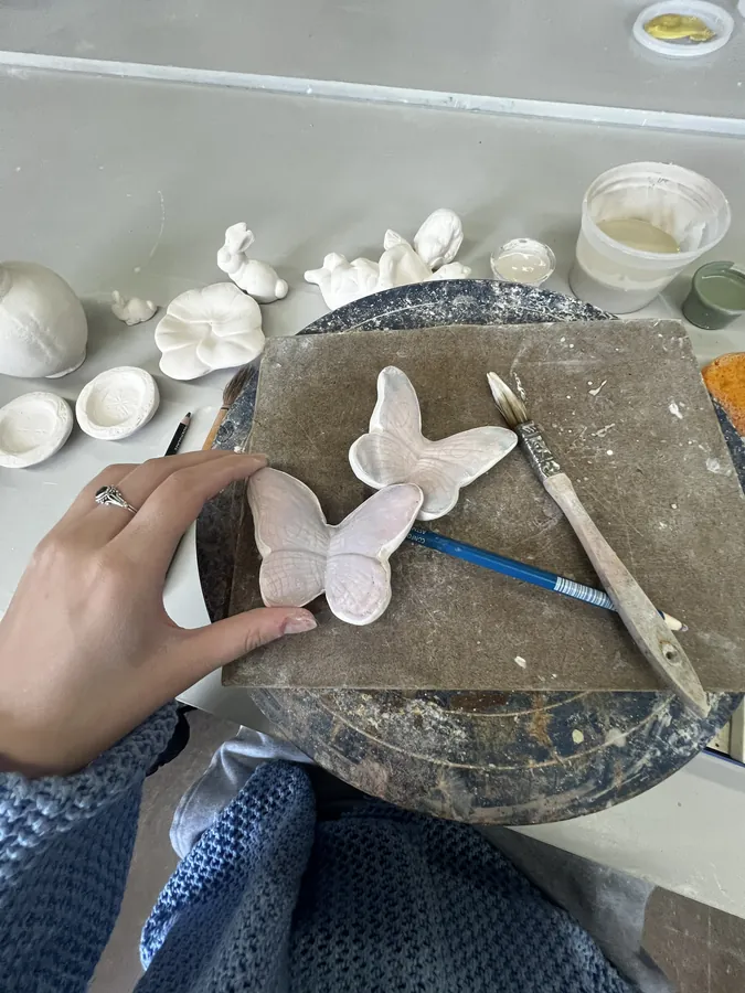 Hands shaping small white clay butterflies on a slab, with sculpting tools alongside.